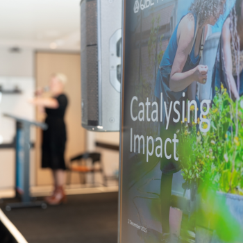 A "Catalysing Impact" event sign in the foreground with a speaker presenting at a podium in a softly lit venue behind